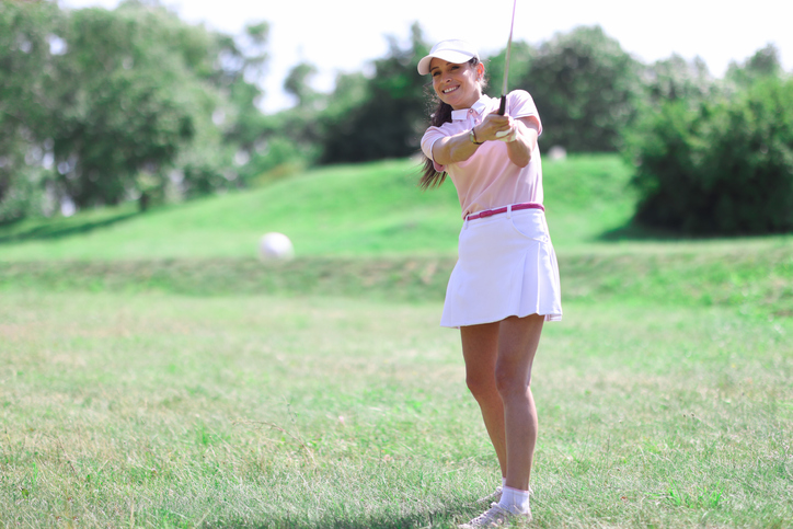 Beautiful woman in white skirt, cap and pink t-shirt play golf on green lawn.